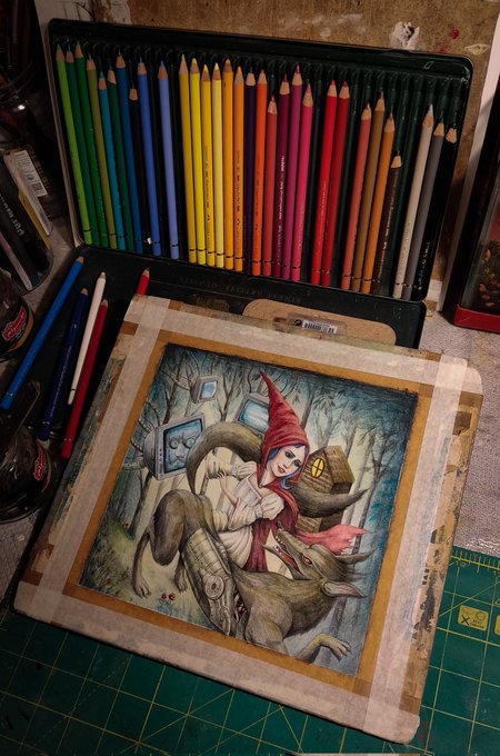 An open tin of colored pencils sits above a detailed illustration of a small red-hooded figure standing on a wooden carving, taped to a cutting mat.