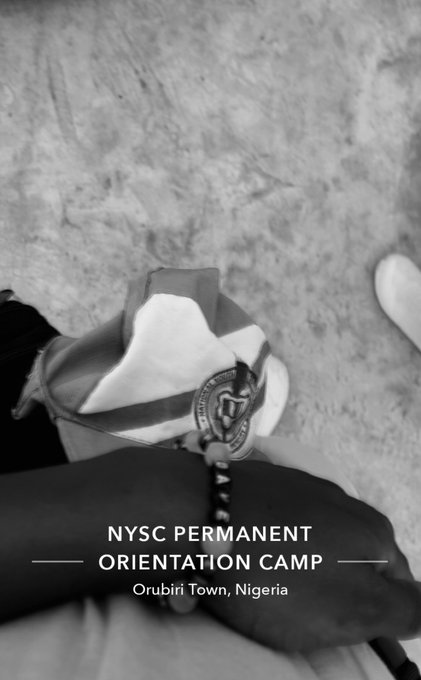 The image features a person wearing a bracelet that says "NYSC Permanent Orientation Camp" written on it. The person's hand is holding a camera, which they are using to take a picture of themselves with the bracelet displayed prominently. The background of the photo appears to be a floor or a wall, but there isn't enough information provided about the specific location or event taking place in the image.