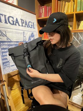 A woman wearing a black cap with a blue logo and a black t-shirt is sitting and holding up a black "Bitget Wallet" backpack, with a banner displaying "TIGA PAGI" and bookshelves visible in the background. The post highlights her showcasing the Bitget Wallet merchandise, aligning with her stylish promotional fit.