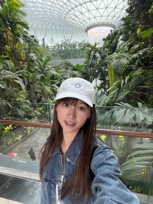 A woman in a white cap and denim jacket takes a selfie at the Jewel Changi Airport, with the iconic Rain Vortex and lush greenery of the Shiseido Forest Valley in the background, capturing a memorable "one last tourist photo" before heading off with her Monad fam.