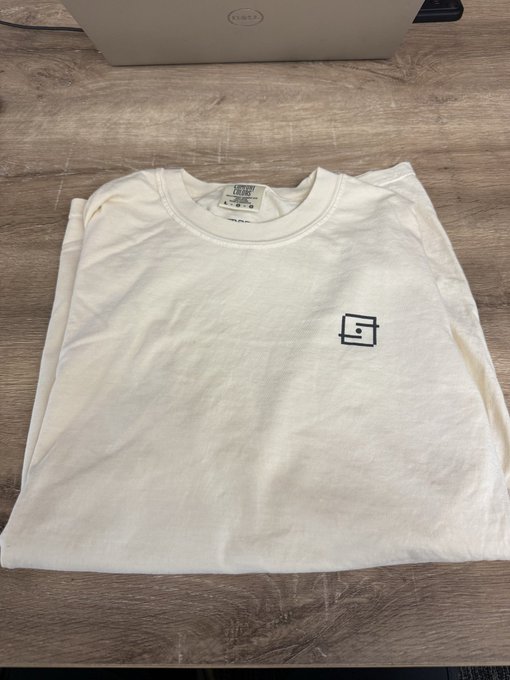 A folded cream-colored Colosseum t-shirt with the brand's logo sits on a wooden table, with a laptop visible in the background—merchandise acquired ahead of a presentation at the MetaDAO Project ownership meeting.