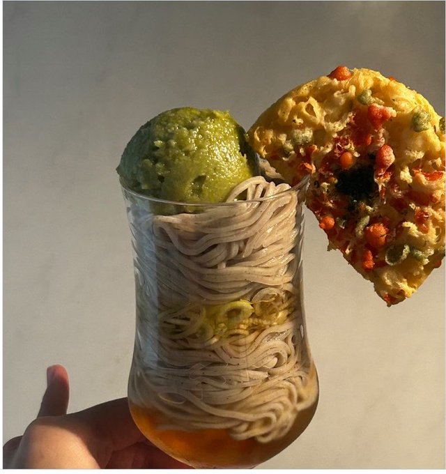 A unique food presentation in a vase-shaped glass featuring layered noodles, broth, lime, guacamole, and a nacho chip with toppings.