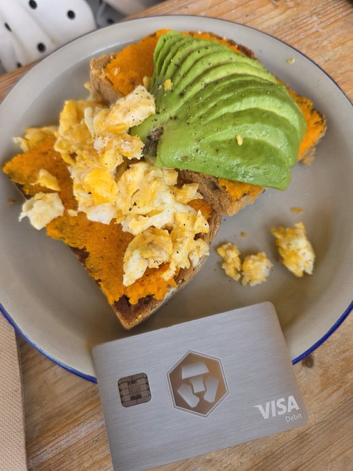 The image features a plate of food consisting of avocado toast, scrambled eggs, and possibly some other ingredients such as bacon or cheese. The plate is placed on top of a wooden table. There are also two pieces of paper present in the scene - one is a credit card with a hexagonal shape and the other appears to be a Visa card.