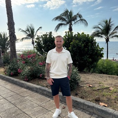 A photo of a smiling man with tattoos on his arms wearing a white polo shirt and black shorts, standing on a paved walkway near palm trees and tropical foliage with the ocean visible in the background.