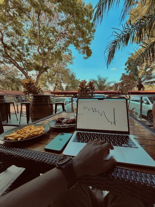 A person is working on a laptop displaying a stock chart at an outdoor restaurant, with a plate of fries and steak visible on the table.