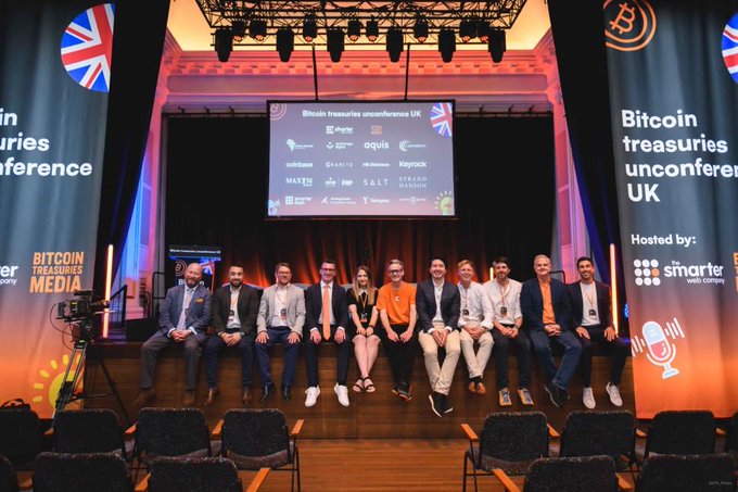 The image shows a group of men sitting on chairs, posing for a picture at an event with a large screen displaying logos related to Bitcoin and Treasures. The event appears to be focused on these digital currencies, as evidenced by the text on display in the background. The individuals seated together suggest that this could be a gathering or conference where attendees have come to discuss and learn about the growth of Bitcoin and its implications for traditional financial systems.