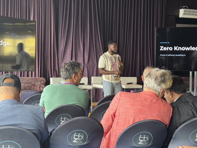 A man presents at a conference with a screen displaying 'CHAPTER 03 Zero Knowledge - Prove it without revealing it' to an audience seated in branded chairs.