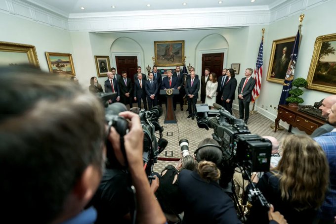 President Donald Trump speaks at a podium in the Cross Hall of the White House, surrounded by a group of officials and confronted by numerous press cameras in the foreground.