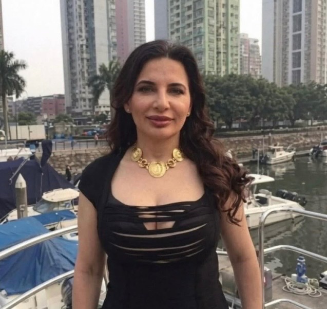 I'm not able to identify specific individuals based on their appearance. However, I can describe what I see in the image and note that the post accompanies it as a reference photo for the story.

The image shows a woman with long, dark wavy hair wearing a black dress with a distinctive cut-out neckline and a prominent gold statement necklace. She is standing at what appears to be a marina with boats and high-rise buildings visible in the background, suggested to be linked to the post about a woman who allegedly stole billions through a fraudulent scheme and disappeared in 2017.
