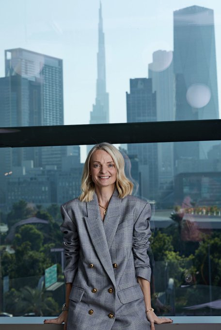A woman with blonde hair wearing a gray double-breasted blazer stands smiling on a balcony with the Dubai skyline, including the Burj Khalifa, visible behind her.