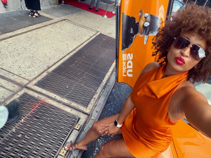 A woman in an orange dress and sunglasses takes a selfie in front of an orange backdrop with partial text reading 'als' and 'nde'.