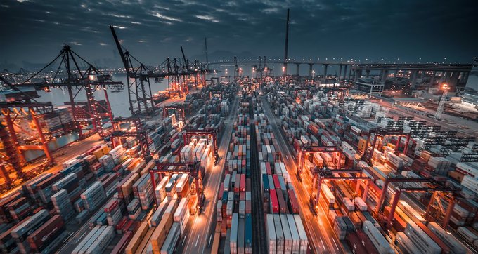 An aerial nighttime view captures a massive, illuminated shipping container port with cranes and a bridge in the background under a cloudy sky.
