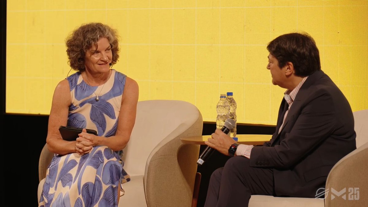 A woman in a blue floral dress sits in a beige armchair, smiling and holding a small device, while a man in a dark suit holds a microphone, seated across from her in what appears to be a panel discussion on a stage with a bright yellow backdrop. Two water bottles sit on a small table between them, and a "AM25" logo is visible in the bottom right corner, connecting to the post's reference to Meridian 2025.