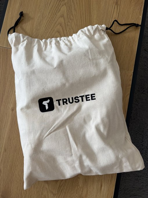 A white drawstring bag rests on a wooden floor, featuring the black "TRUSTEE" logo alongside a gun icon, which appears to be branded merchandise received from Trustee.