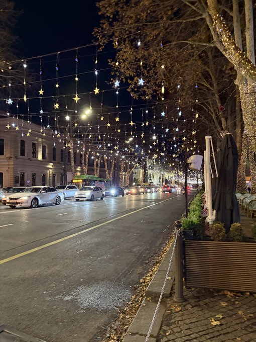 A nighttime city street is illuminated by rows of hanging star-shaped string lights stretching over the road, with parked cars lining the curb and a closed patio umbrella visible in the foreground. The festive, festive atmosphere suggests a seasonal celebration, fitting the casual "See you later" sign-off from the GN web3 post.