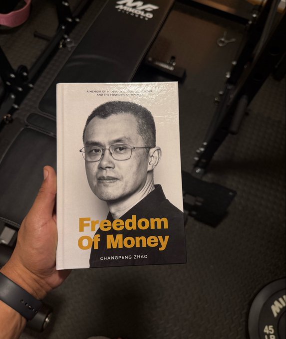 A hand holds up Changpeng Zhao's memoir "Freedom Of Money" — a memoir of #CZBT[CEO] and the founding of @Binance — in front of home gym equipment, tying together the post's workout session with the arrival of CZ's newly released book.