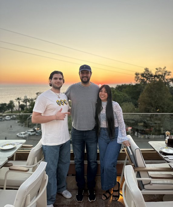 Three people pose together on a rooftop terrace at sunset, with one wearing a white t-shirt reading "GOAT," a striped shirt, and a lace top, while the caption hints at an upcoming international trip with social media personalities @CryptaCobra and @Lunaticanto. The scenic ocean backdrop and warm golden sky suggest a Mediterranean destination, tying into the mystery travel reveal.