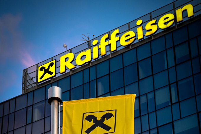 The exterior of a Raiffeisen bank building is shown, featuring its logo and yellow flag against a blue sky.