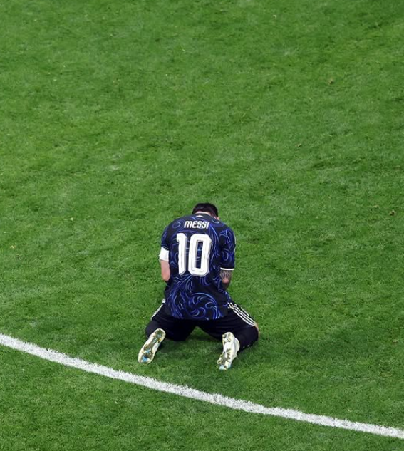 An aerial photo of Lionel Messi wearing the number 10 Argentina jersey, kneeling on the grass field with his head bowed down.
