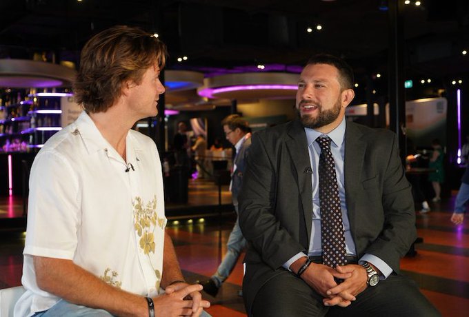 Two men are seated and conversing in what appears to be an interview setting; the man on the right is wearing a dark suit and tie, while the man on the left wears a white patterned shirt, with a dimly lit bar or lounge visible in the background.