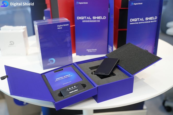 A photograph of a Digital Shield product display, featuring blue packaging boxes branded 'DIGITAL SHIELD' with a smartphone and security device resting inside an open presentation case.