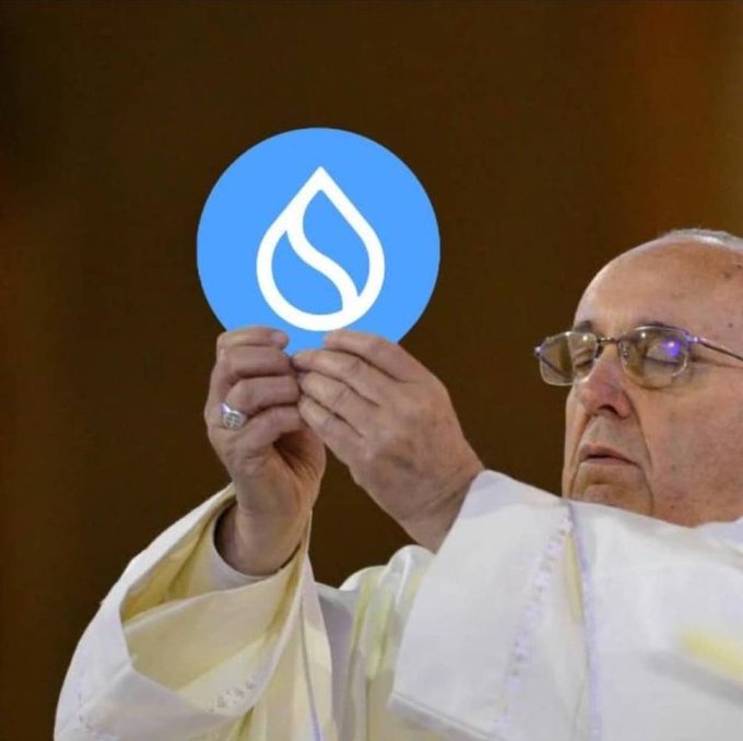 A man wearing white religious vestments and glasses holding up a Sui (SUI) cryptocurrency logo on a blue circular sign.