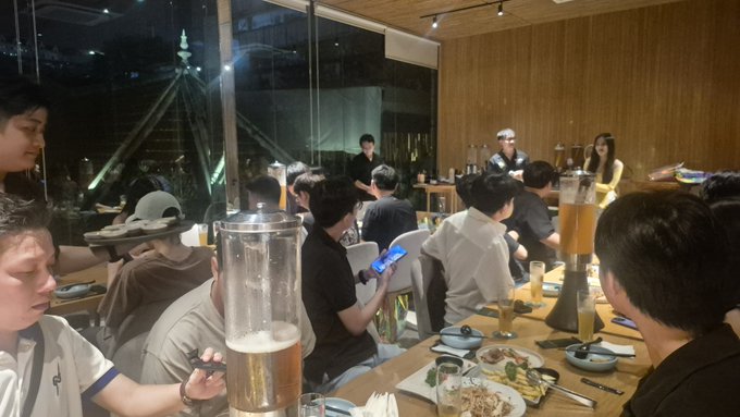 A busy restaurant interior at night with diners seated at tables, a server carrying plates, and a large window showing a bridge outside.