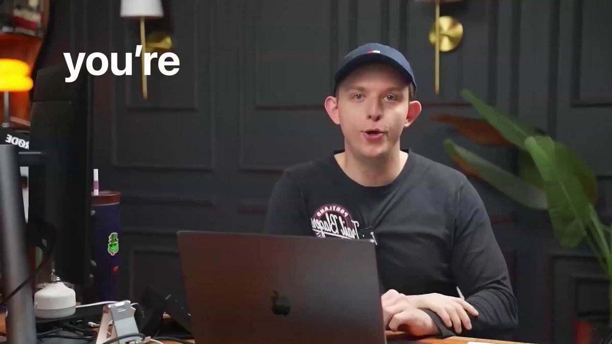 The image features a man sitting at a desk with an open laptop computer, wearing a black baseball cap. The background of the image shows a dark room or office setting, which complements the overall theme of the photo. The caption "you're" is written in white text above the man's head and mouth, suggesting that he might be speaking into the microphone present in the scene.