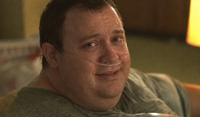 A close-up of a middle-aged man with an oxygen tube in his nose, appearing tearful, set in a dimly lit room with a lamp visible in the background.