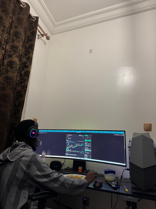 A young man wearing glowing headphones and a plaid hoodie sits at a desk focused on an ultrawide monitor displaying a candlestick crypto trading chart, with a plate of food beside him—capturing the late-night grind of a degen crypto trader scanning the markets for plays.