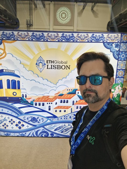 A selfie of a man wearing sunglasses and a lanyard standing in front of an 'ETHGlobal Lisbon' event backdrop featuring colorful illustrations of Lisbon landmarks.