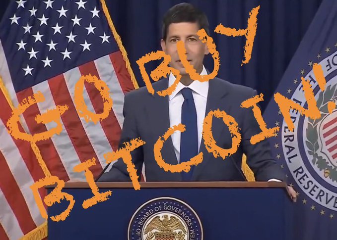 A man in a suit speaks at a podium with American flags and Federal Reserve seal behind him, with orange graffiti-style text reading 'BOY BITCH' overlaid on the image.