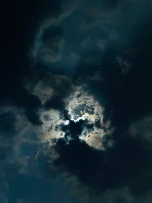 A moody sky with dark clouds partially obscuring a glowing moon, with moonlight filtering through the cloud cover.