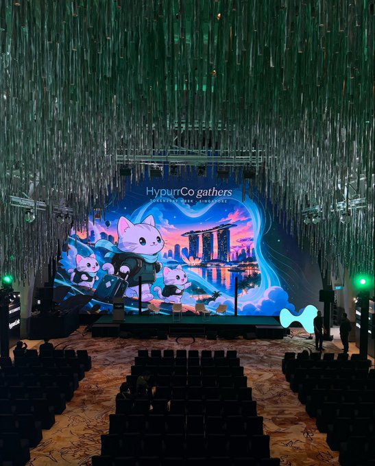 Empty auditorium with rows of chairs facing a stage featuring a 'Hypurr Co gathers' Singapore event backdrop with animated cat characters and Marina Bay Sands imagery.