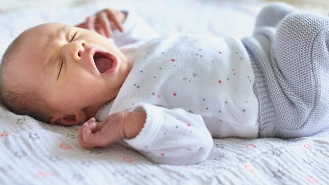 A newborn baby yawns while lying on a star-patterned blanket, wearing a white long-sleeved top and gray knitted pants.