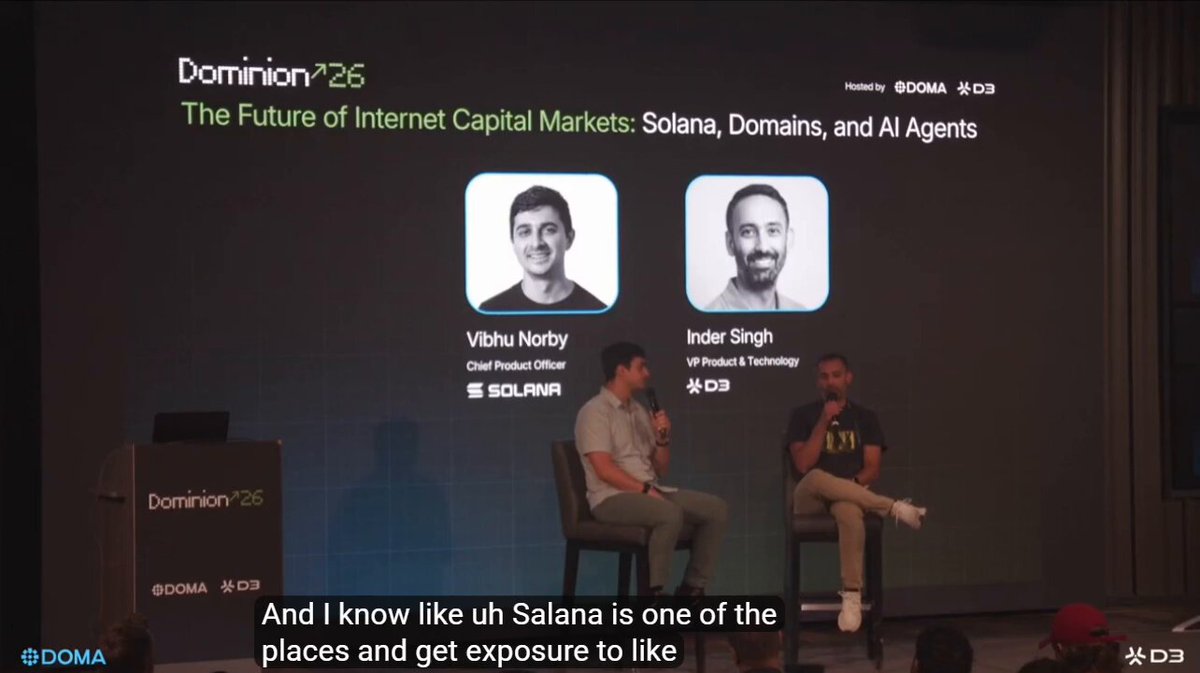 The image shows a stage where two men are speaking about Internet Capital Markets. They both sit on chairs behind a podium as they discuss these markets. The backdrop features a black wall with blue text indicating that "Domain capital markets - What most are missing Doma = where the domain becomes an onchain asset Solana = where this asset can find traders, liquidity, LPs, wallets, and DeFi activity @domaprotocol 's own protocol will bridge DNS infrastructure with ecosystems like Solana". The speakers appear to be presenting information about these markets.