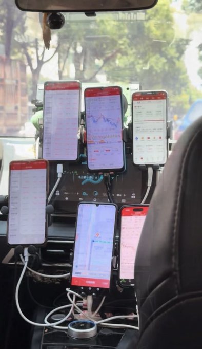 A view inside a vehicle showing a multi-phone mount holding six smartphones displaying various apps and charts, with a person's shoulder visible on the right.