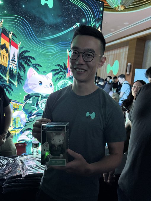 A man wearing a gray t-shirt with a green icon smiles and displays a labeled "MYPURRR" collectible box, standing in front of a vibrant promotional backdrop featuring cartoon cats and palm trees. The image ties to the "Hyperliquid" post as part of an iconic photo opportunity at what looks like a crypto/NFT brand activation event.