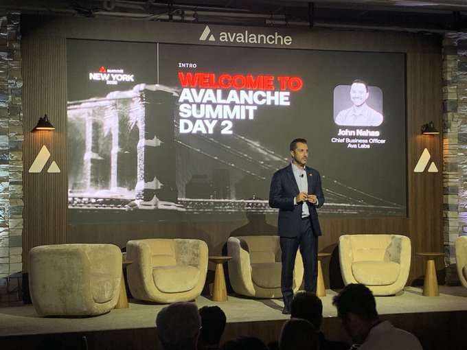 John Nahas, Chief Business Officer of Ava Labs, stands on stage delivering the welcome address at "Avalanche Summit Day 2" in New York, as displayed on the screen behind him. The event, hosted by Ava Labs, features comfortable lounge seating on stage as he addresses the audience.