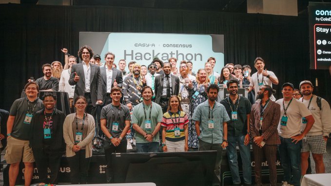 A group photo of participants and organizers at an "EASY-A CONSENSUS Hackathon" event, with attendees wearing lanyards and posing on stage.