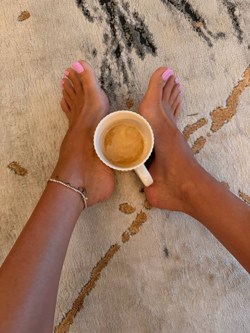 Bare feet with bright pink toenail polish rest on a patterned rug while holding a small mug of espresso, perfectly capturing the cozy, self-care moment the caption alludes to.
