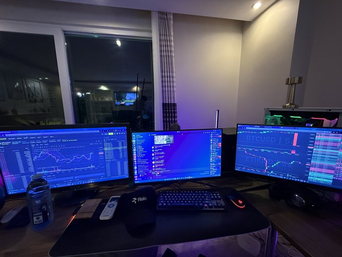 A multi-monitor trading setup displays BTCUSD and ETHUSD charts alongside chat windows, with a microphone labeled "Rialo" on the desk. The image visually parallels the post's message about @RialoHQ bridging Solana SVM compatibility with broader blockchain flexibility, reflecting the dual-environment workflow the project promotes.