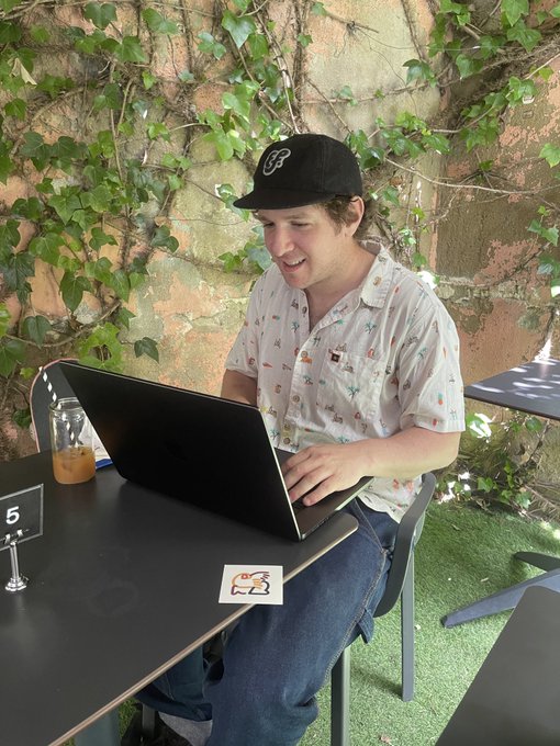 The image features a man sitting at an outdoor table, wearing a baseball cap and using his laptop computer. The background of the scene includes green plants and trees, creating a pleasant atmosphere for working or studying outdoors. The caption "Mint day with this guy. LFG! @EfdotStudio" suggests that the man is enjoying his time outside while working on his laptop.
