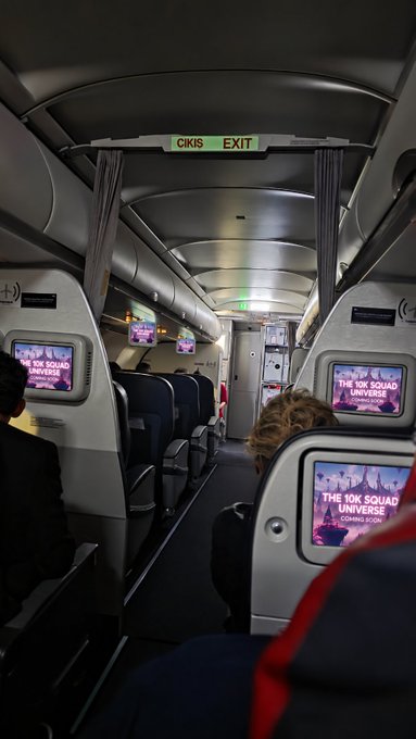 The interior cabin of an airplane viewed from a passenger seat, showing screens displaying 'THE 10K SQUAD UNIVERSE' and a green illuminated 'CIKIS EXIT' sign above the aisle.