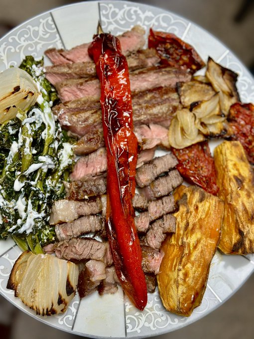 A plate is filled with sliced grilled meat, a whole roasted red pepper, roasted onions, a creamy green spinach dish, roasted tomatoes, and grilled eggplant.