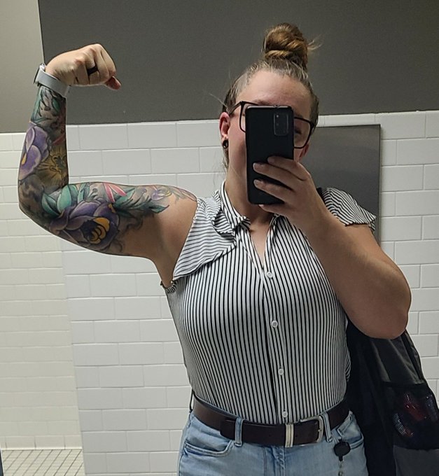 A mirror selfie shows a person with a floral tattoo sleeve flexing their arm, wearing a striped sleeveless top and jeans, in a tiled bathroom.