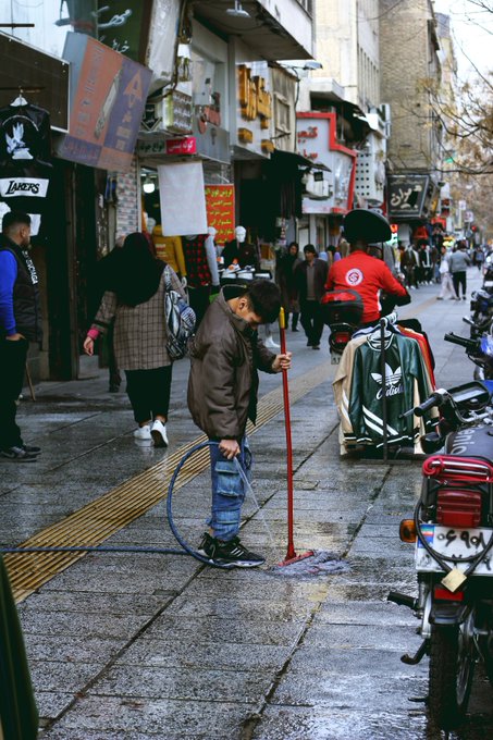 A young man in a brown jacket uses a hose to clean a stone sidewalk in a busy street, with motorcycles parked nearby and Persian/Farsi signage visible on the buildings.