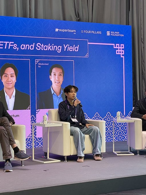 A young man sits in a white chair on stage speaking into a microphone at a panel discussion, with a blue backdrop displaying 'superteam VN', 'FOUR PILLARS', 'SOLANA FOUNDATION', and partial text reading 'ETFs, and Staking Yield'.