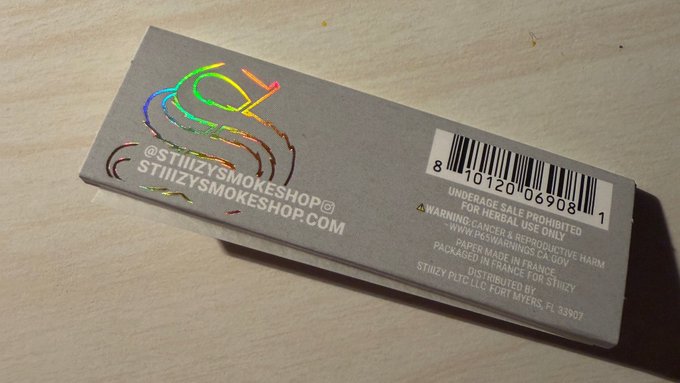 The back of a gray product box with a holographic sticker from @STIIIZYSMOKESHOP with a barcode and warning label.
