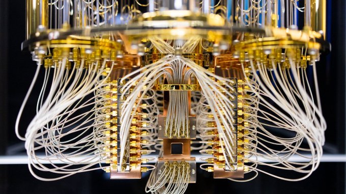 Close-up photograph of a quantum computer or advanced technological device featuring intricate gold wiring and components suspended in a cylindrical chamber.